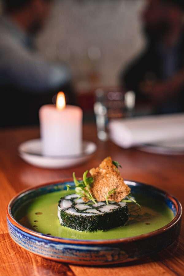 A plated gourmet dish featuring a circular seaweed-wrapped roll in a green sauce, garnished with microgreens and a crispy element, served beside a lit white candle.