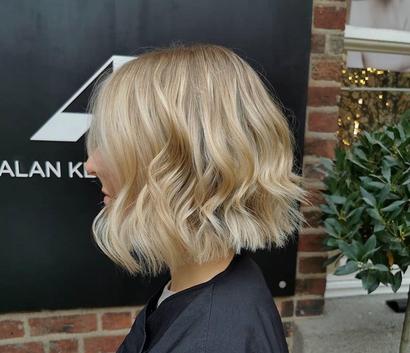 Woman with a blonde, shoulder-length wavy bob haircut. The hair has subtle layers and soft beachy waves, with a natural blend of warm and cool tones. She is photographed in profile against a black salon sign and a brick wall, wearing a dark salon cape.
