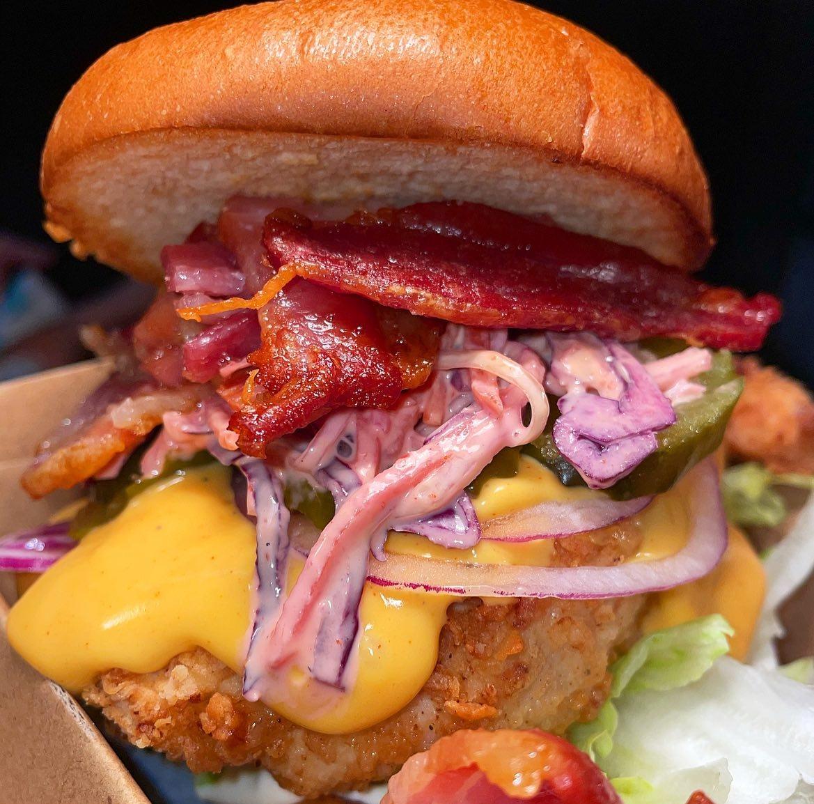 Close-up of a fried chicken sandwich with crispy bacon, creamy coleslaw, pickles, sliced red onion, melted yellow cheese, and lettuce, all inside a toasted bun.