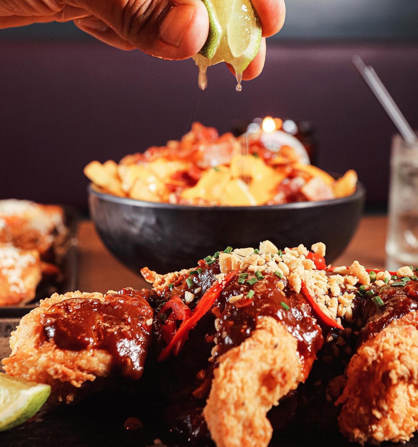 Close-up of lime juice being squeezed onto fried chicken topped with dark sauce, chopped peanuts, and sliced red chili, with cheesy fries in the background.