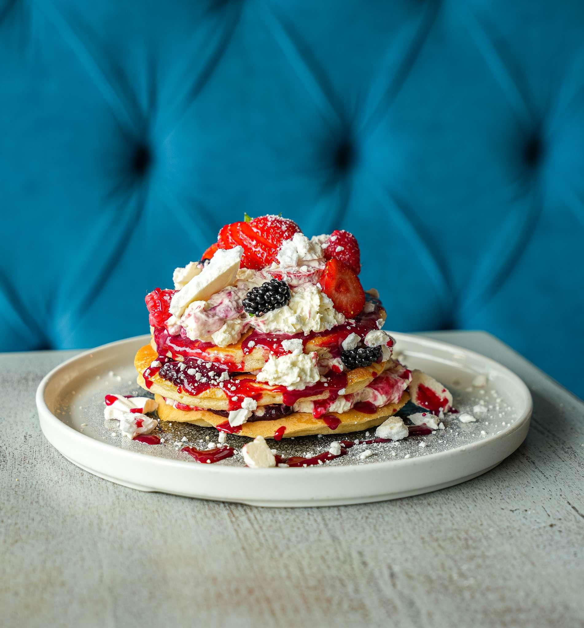 Stack of golden pancakes topped with whipped cream, strawberries, blackberries, berry sauce, and crushed meringue on a white plate.
